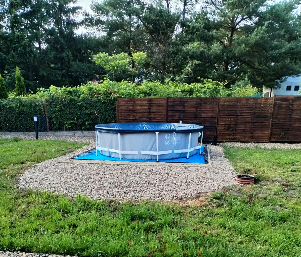Round garden pool on a gravel island by the wooden fence, pine trees in the background