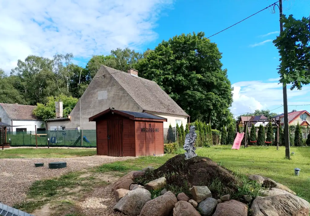Outbuilding with the sign “11 Wrzosowo”, a rockery with a bird figure and a playground with swings and a slide on the lawn