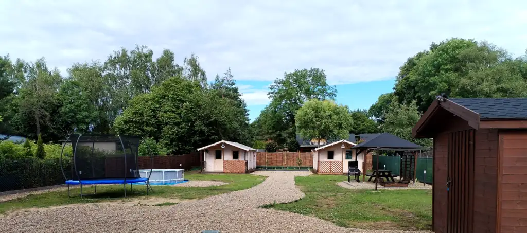 Szeroki widok na ogrodzony teren: trampolina, basen ogrodowy, domki letniskowe, altana i drewniany budynek gospodarczy